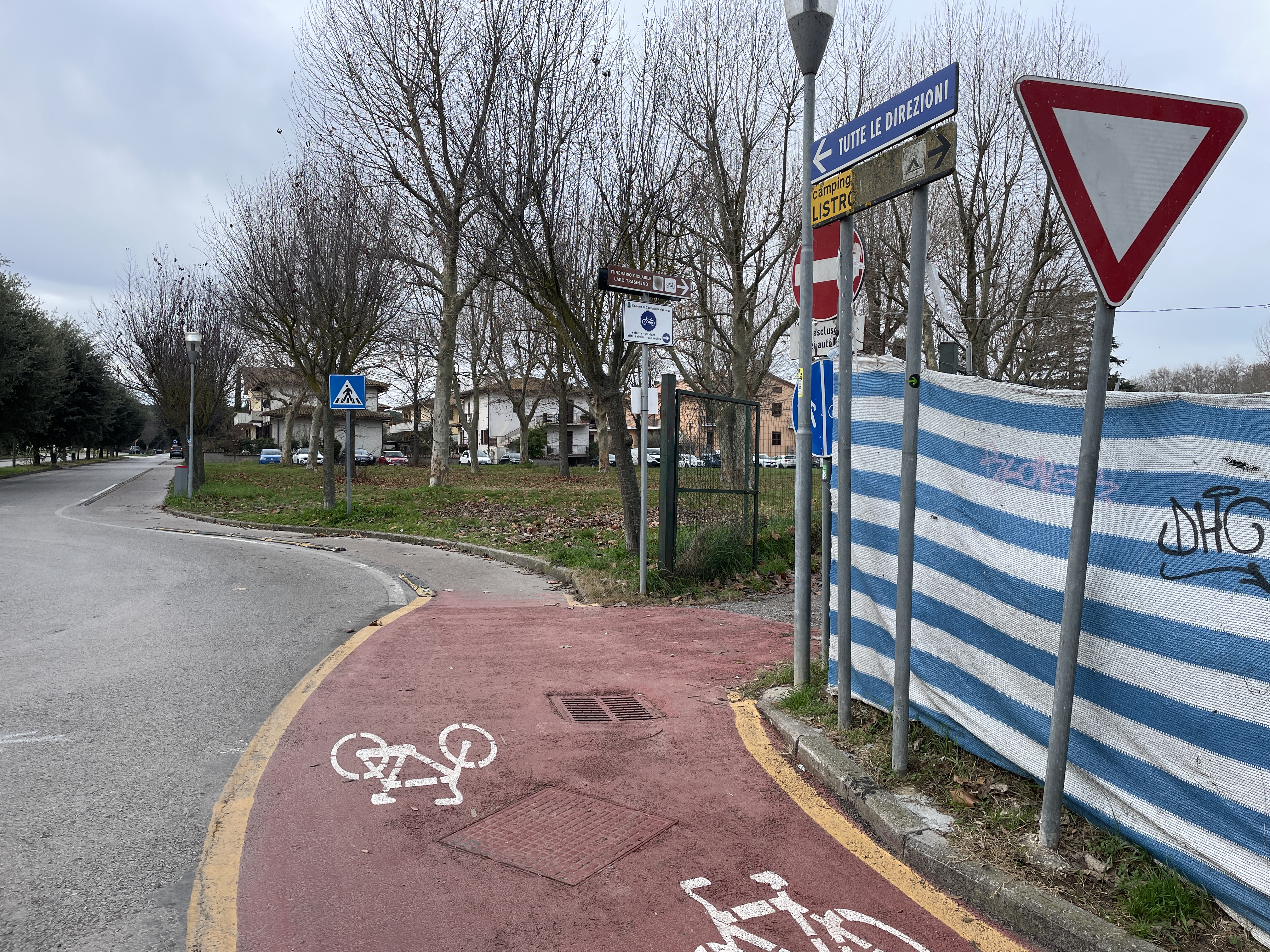 Tratto di ciclovia su pista rossa che gira a destra a fianco di una strada asfaltata. Sempre sul lato destro, vari segnali stradali